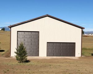 A light-colored metal building with two brown garage doors, one tall and one wide, stands on a dry, grassy lot with a small tree in front.
