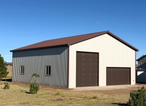 A metal building with a brown roof, one tall garage door, one standard garage door, and two windows, set on a grassy lot under a clear blue sky.