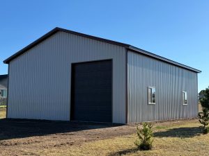 A large metal storage building with a dark roll-up door and two windows stands on a grassy lot under a clear blue sky.