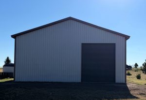 A large metal building with a dark garage door, standing on a grassy plot under a clear blue sky.