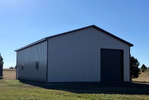 A large metal storage building with a peaked roof and a closed roll-up door stands on grassy land under a clear blue sky.