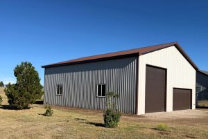 A metal building with two brown garage doors and two windows stands on a grassy lot under a clear blue sky.