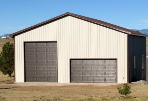 A beige metal building with two brown garage doors, one tall and one standard size, sits on a gravel patch in a grassy field under a clear blue sky.