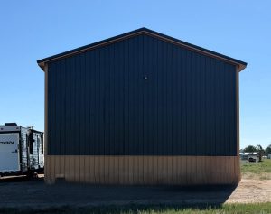 A large, blue and tan metal building stands on dirt, with an RV partially visible on the left side under clear blue skies.