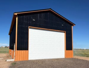 A large black and orange metal building with a tall white garage door, set on a gravel surface under a clear blue sky.