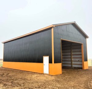 A large black and orange metal barn with a white door and a tall open entry, standing on bare soil with a foggy sky in the background.