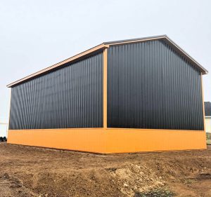 Large metal building with black siding and orange trim, situated on bare ground under a cloudy sky.